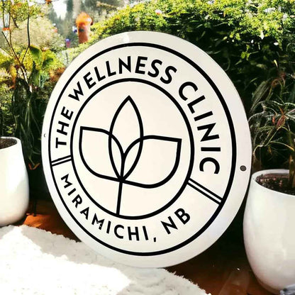 The Wellness Clinic Miramichi, NB circular sign with leaf logo, displayed outdoors among greenery.