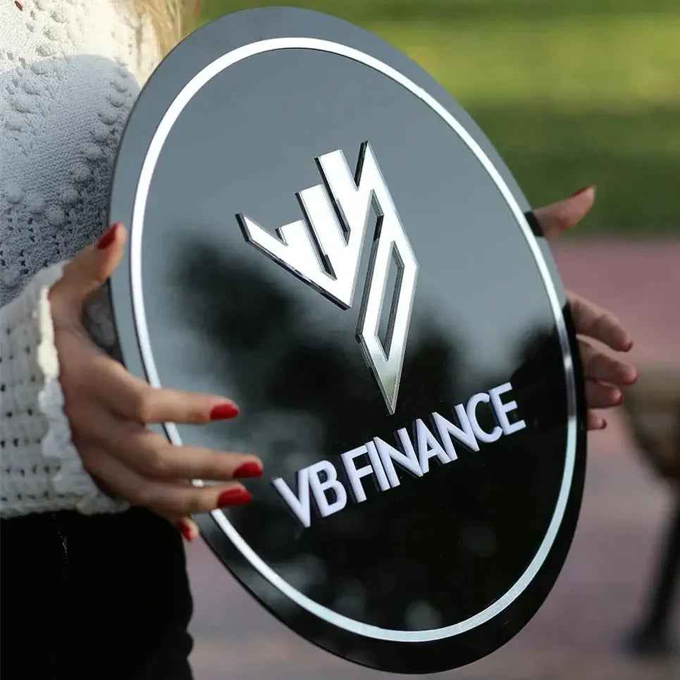 Person holding black oval VB Finance sign with silver logo and white border