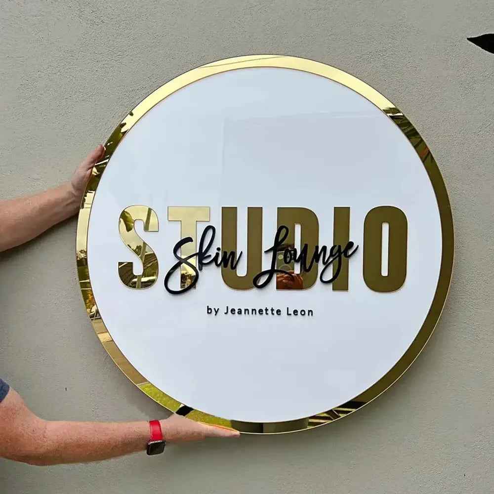 Round gold-framed sign with 'STUDIO Skin Lounge by Jeannette Leon' in elegant lettering, held against wall