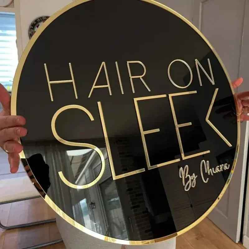 Circular black and gold neon-style sign reading 'HAIR ON SLEEK By Chiara', held indoors